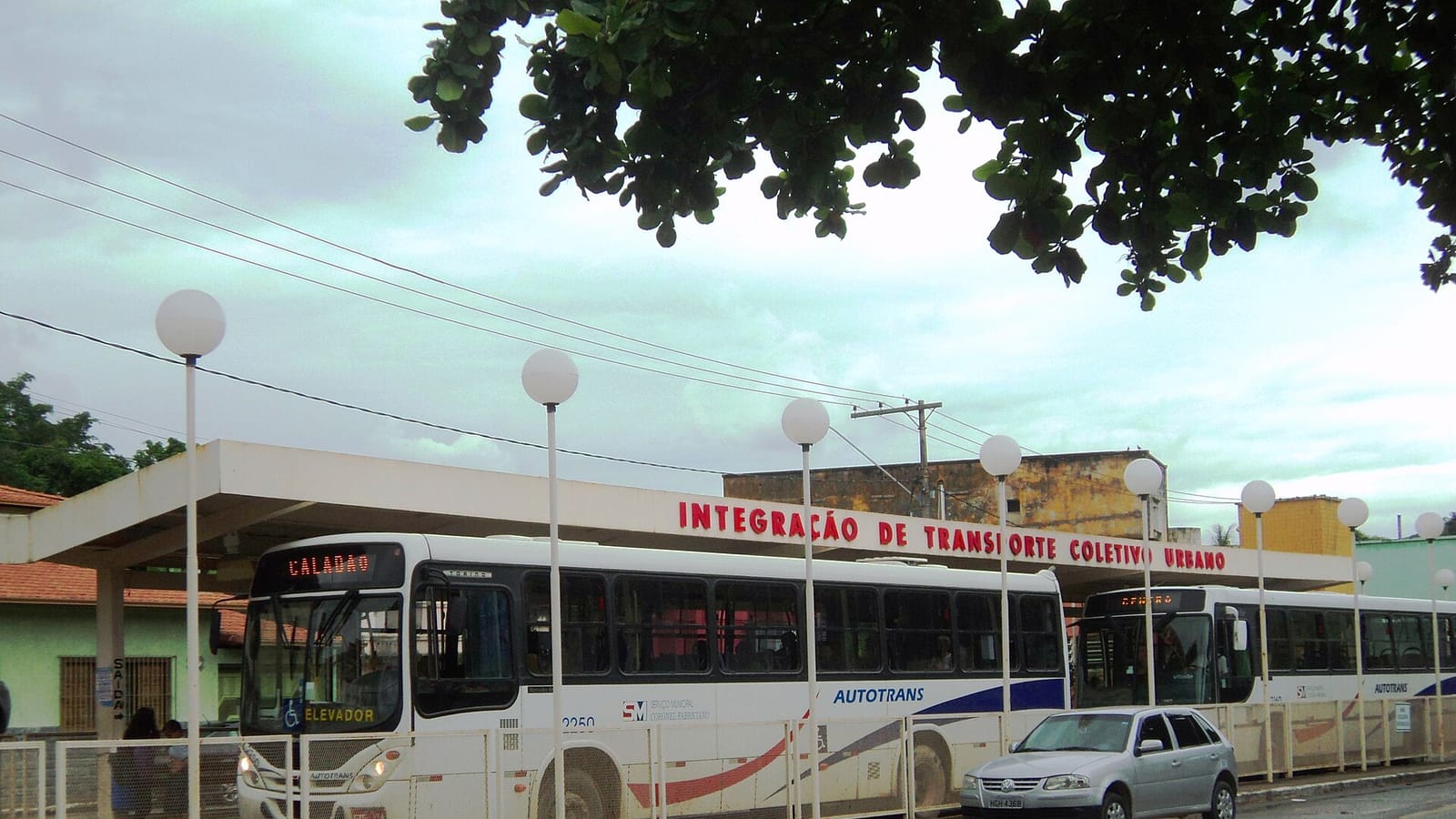 Ônibus do transporte coletivo urbano circulando por uma rua da cidade, com passageiros esperando na parada